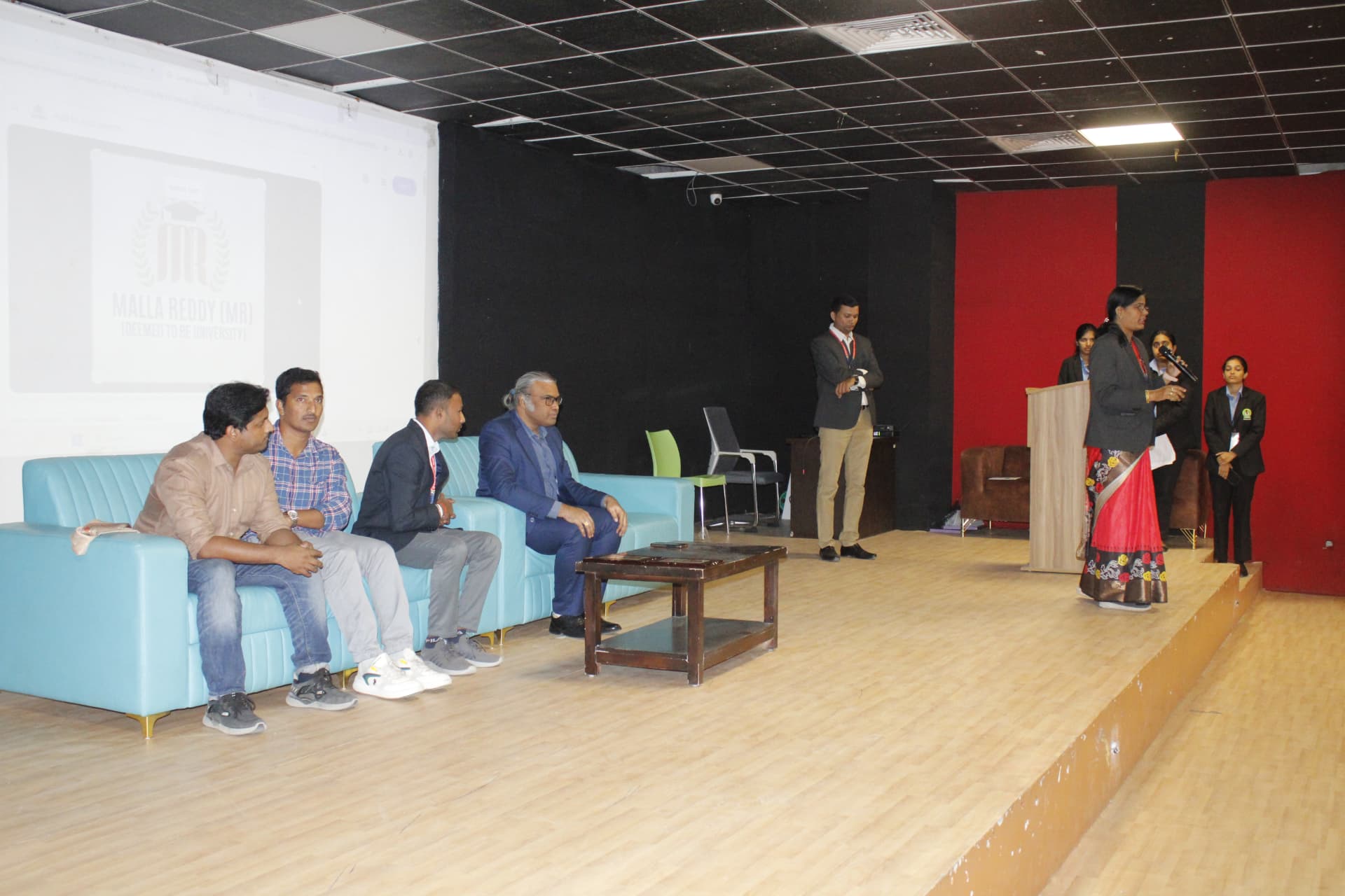 An event speaker addressing the auditorium with Navaneeth Reddy, Velamarthy Gopichand, and college representatives seated on stage